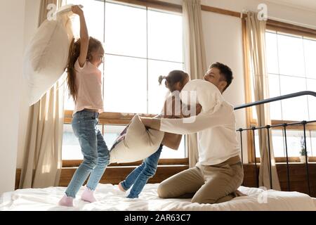 Il cuscino di un giovane padre con gioia combatte con le figlie in camera da letto Foto Stock