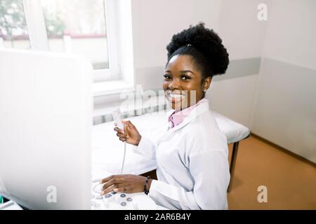 Giovane e allegro operatore donna africana di una macchina di scansione a ultrasuoni che analizza i risultati diagnostici del paziente. Giovane medico africano sorridente che lavora Foto Stock