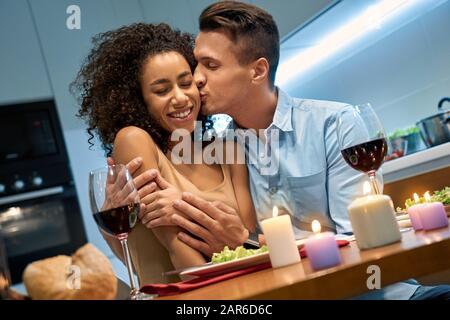 Data Romantica. Giovane coppia multietnica a casa seduta a tavola cena abbracciando fidanzato abbracciando ragazza baciare la sua sorridente allegro Foto Stock