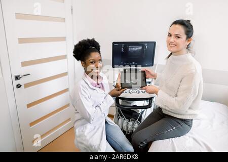 Donna incinta caucasica felice visita medico presso ospedale o clinica medica per consulente di gravidanza. Donna africana ginecologista con paziente tiene il Foto Stock