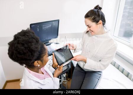 Giovane donna caucasica incinta e medico africano che guarda insieme l'immagine ecografica del bambino, tenendo la compressa digitale Foto Stock