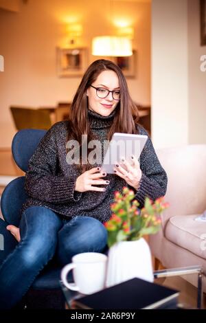 Scatto ad alto angolo di giovane donna sorridente che si rilassa in poltrona a casa mentre usa il tavolo digitale e naviga in rete. Foto Stock
