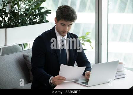 Giovane uomo d'affari esperto che analizza la documentazione finanziaria nel moderno posto di lavoro. Foto Stock