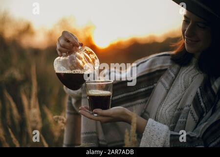 Donna hippster in cappello versando caffè caldo fresco in tazza di vetro sullo sfondo di luce calda soleggiata in erbe rurali. Caffè alternativo in viaggio. Atm Foto Stock