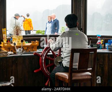 Uomo al volante del capitano in nave da crociera con i turisti che scattano foto sul ponte, Halong Bay, Vietnam, Asia Foto Stock