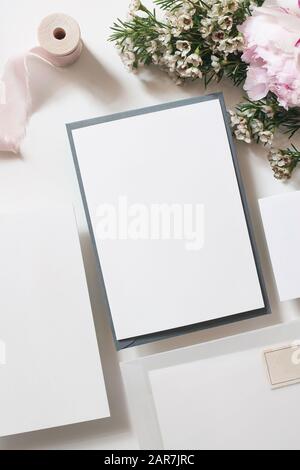 Matrimonio, cartoleria compleanno simulazione scena. Biglietti d'auguri bianchi, busta vellum e francobollo. Composizione forale di peonie rosa, cameleucium f Foto Stock