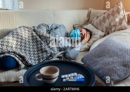 Donna malata che ha influenza o freddo. Ragazza sdraiata a letto indossando maschera protettiva da pillole e acqua sul tavolo Foto Stock