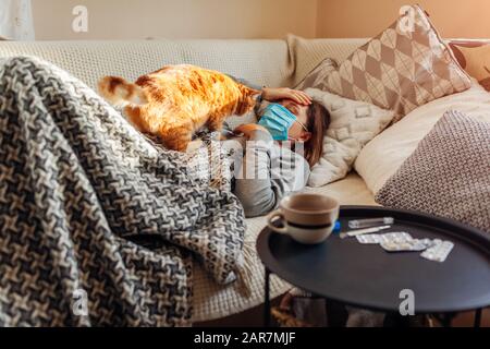 Donna malata che ha influenza o freddo. Ragazza sdraiata a letto con gatto indossare maschera protettiva da pillole e acqua sul tavolo Foto Stock