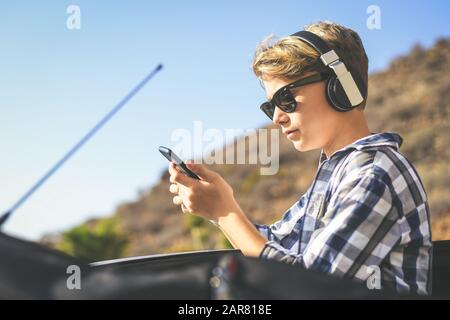Ritratto di un quartiere alla moda giovane ragazzo in ascolto di musica con il telefono cellulare proveniente dal tetto aperto di un'auto. Teen godendo di una splendida giornata di primavera per esterno Foto Stock