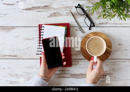 Ambiente di lavoro moderno con la mano della donna che usa il telefono cellulare e beve caffè. Vista dall'alto. Disposizione piatta. Foto Stock