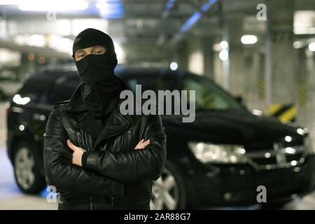 Criminale maschile in giacca nera e balaclava in piedi sul parcheggio Foto Stock