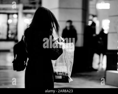 Una figura offuscata e irriconoscibile di una giovane ragazza che si scagliava giù per strada con un cappotto scuro e un telefono cellulare nelle sue mani. Silhouette stilizzata di un giovane moderno Foto Stock