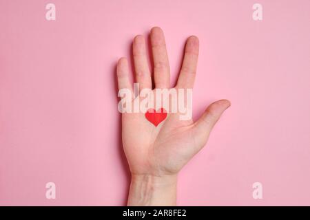 Nel palmo della mano si trova un piccolo cuore rosso su uno sfondo di carta rosa. Vista dall'alto. Foto Stock
