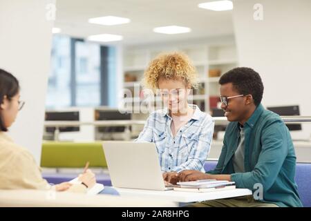 Gruppo multietnico di studenti che usano il computer portatile e sorridono felicemente mentre lavorano al progetto nella biblioteca universitaria, copia spazio Foto Stock