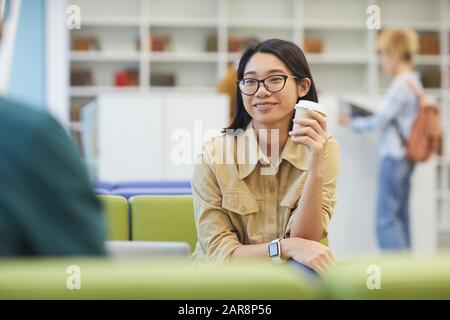 Ritratto di ragazza asiatica sorridente godendo del caffè mentre studia nella biblioteca dell'università, copia lo spazio Foto Stock