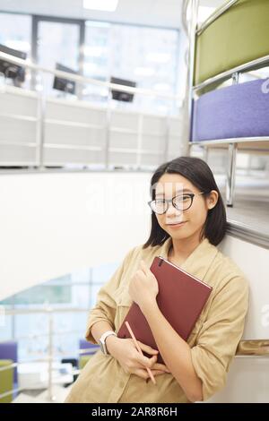 Girovita u ritratto di giovane donna asiatica sorridente alla macchina fotografica mentre in piedi da scala a spirale in moderno college interno, copia spazio Foto Stock