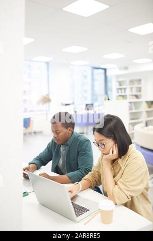 Ritratto ad alto angolo di due studenti che studiano nella biblioteca universitaria, fuoco sulla giovane donna asiatica che usa il laptop in primo piano, spazio della copia Foto Stock