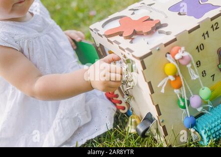 Carino bambina sta giocando con busiboard all'aperto sull'erba verde. Giocattoli educativi per i bimbi. ragazza porta aperta a cubo del bordo Foto Stock