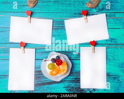 Foglio quadrato bianco di carta è attaccato da clothespins di legno rosso nella forma di cuori su superficie di legno blu chiaro, con torta di Pavlova deliziosa con Foto Stock