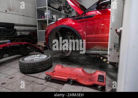 Vettura usata rossa con cofano aperto sollevato su un ponte elevatore per riparare il telaio e il motore con le ruote rimosse e il paraurti rotto in un'officina di riparazione del veicolo. A. Foto Stock