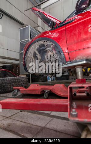 Vettura usata rossa con cofano aperto sollevato su un ponte elevatore per riparare il telaio e il motore con le ruote rimosse e il paraurti rotto in un'officina di riparazione del veicolo. A. Foto Stock