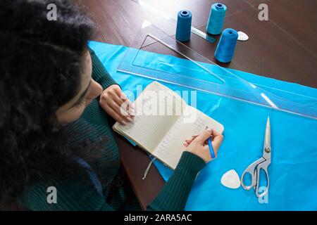 bella seamstress con lunghi capelli scrive in un notebook. il sarto crea una collezione di abiti. giovane donna designer vestiti note idee in nota Foto Stock