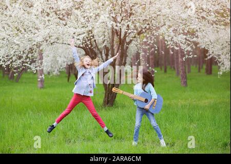 concerto di musica rock estatica dal vivo all'aperto di due ragazze piccole che saltano cantando e suonando la chitarra nel verde parco primaverile dei fiori Foto Stock