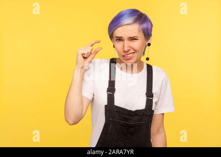 Ritratto di giovane ragazza hippster con viola capelli corti in tute di denim che mostrano un piccolo gesto dito, poco segno di punta e guardando la fotocamera con Foto Stock