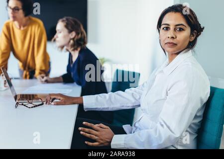 Fiducioso giovane donna seduta a una presentazione aziendale con i colleghi in sala riunioni. Femmina con i colleghi in sala conferenze guardando la fotocamera. Foto Stock