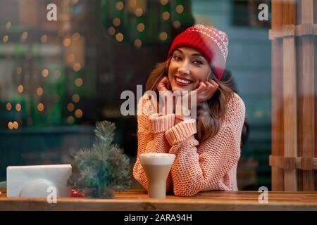 Bella donna bionda caucasica in cappello a maglia rossa seduto dietro la finestra della caffetteria bevendo una calda latte in un meraviglioso giorno di Natale. curly europeo Foto Stock