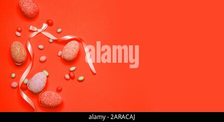 Composizione creativa minimale per le vacanze con decorazioni di uova, fagioli di gelatina dolce, nastro rosa seta su sfondo arancione con copyspace. Biglietto di auguri per buona pasqua, compleanno, giorno delle madri, banner sconto Foto Stock