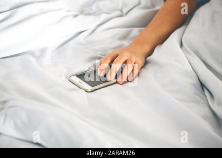 La mano da vicino tiene il telefono a letto. La ragazza si è addormentato con un telefono in mano ed è addicted. Oppure una persona si rivolge a un telefono cellulare per disattivare la sveglia. Foto Stock