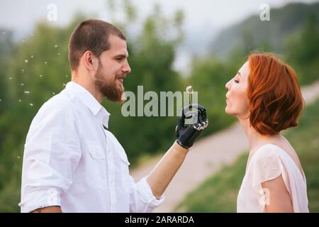 Uomo che tiene il dente di leone in una mano bionica, perché la sua ragazza soffia su di esso. Vista laterale. Foto Stock