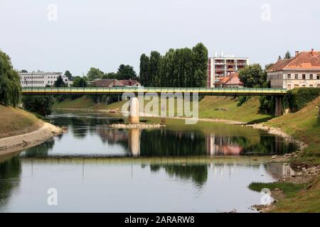 Vecchio ponte di metallo verde e giallo montato su una solida fondazione in cemento sul fiume locale con bassi livelli d'acqua estivi Foto Stock