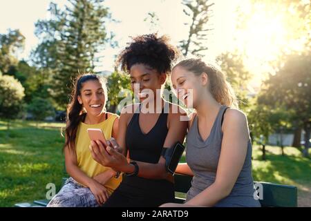 Diversi sportivi giovani amici seduti insieme su panchina guardando il telefono cellulare nel parco al mattino - felice fitness amici sorridenti e. Foto Stock
