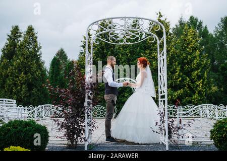 Appena sposata coppia in piedi sotto archi bianchi in metallo in un parco, tenendo le mani Foto Stock