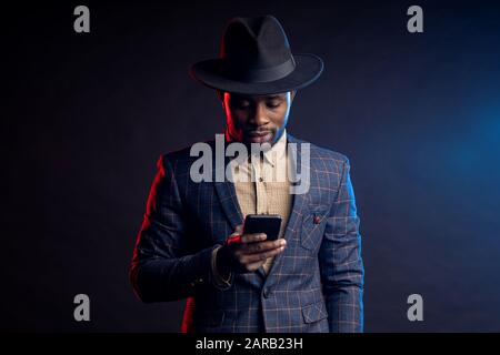Girovita di sicuro elegante ragazzo africano, tenendo il telefono cellulare, guardando lo schermo, leggendo le notizie, chiacchierando con gli amici online, in piedi al buio Foto Stock