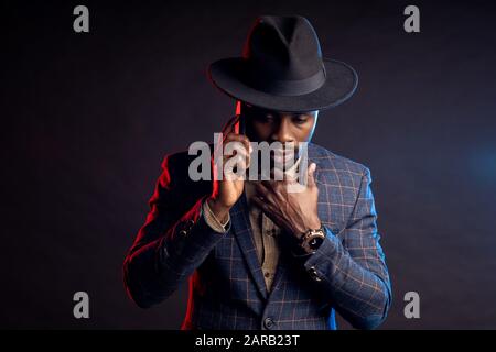 Scatto orizzontale di serio preoccupato giovane africano elegante vestito, cappello, parlando sul telefono cellulare, in piedi in buio su sfondo nero . Suc Foto Stock