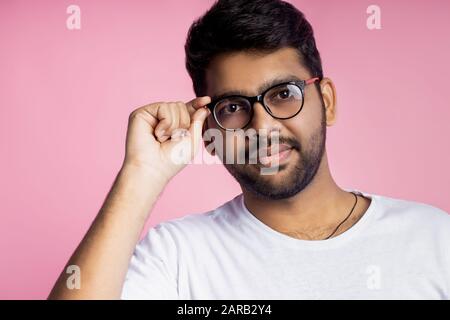 Bel ragazzo indiano intelligente, studente vestito di t shirt bianca, sorridente guardando la macchina fotografica, indossando occhiali e toccandolo, posando in studio su ba rosa Foto Stock