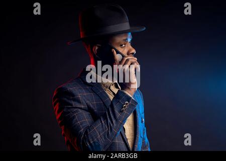 Scatto orizzontale di serio preoccupato giovane africano elegante vestito, cappello, parlando sul telefono cellulare, in piedi in buio su sfondo nero . Suc Foto Stock