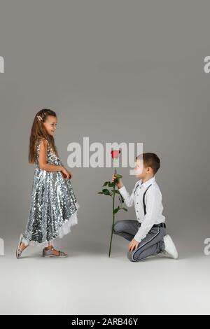 piccolo gentiluomo ragazzo che dà una rosa rossa alla ragazza alla moda bambino in abito d'argento su sfondo grigio. San Valentino. Foto Stock