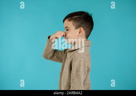 bambino piccolo ragazzo che fa gesto odore cattivo su sfondo blu. espressione facciale. bambino copre il naso con la mano, odore qualcosa di terribile Foto Stock