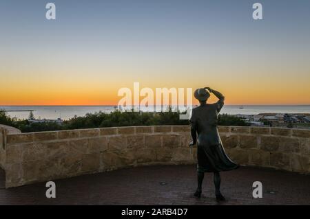 La sera, la statua in bronzo di una donna, con il suo cappello nella brezza marina, si affaccia sul porto di Geraldton presso il monumento commemorativo HMAS Sydney 11. Foto Stock