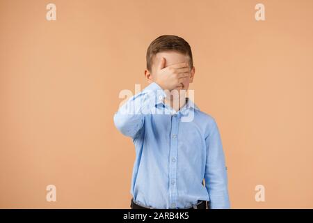 Carino bambino ragazzo chiude gli occhi con la mano su sfondo beige. Foto Stock