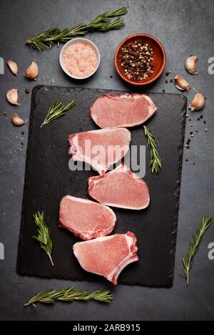 diverse bistecche di maiale crude su una tavola di pietra nera, spezie su sfondo nero. vista dall'alto Foto Stock