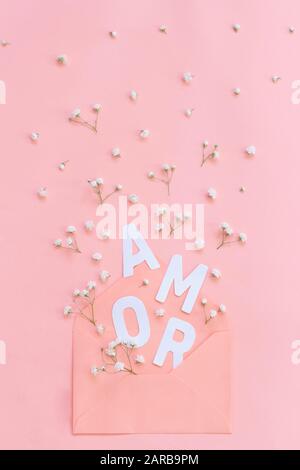 Piccoli fiori bianchi, busta rosa chiaro e parola AMOR su una vista dall'alto di sfondo rosa chiaro Foto Stock