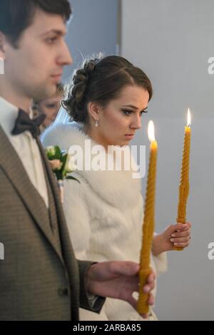 Sposa e sposo in piedi alla cerimonia nuziale. Felice elegante coppia di nozze tenendo candele con luce sotto corone dorate durante il matrimonio santo in Foto Stock