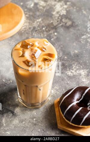 Caffè nero ghiacciato con cubetti di ghiaccio in un bicchiere alto e una ciambella di cioccolato fresco. Bevanda rinfrescante sullo sfondo grigio del tavolo. Messa a fuoco selettiva ravvicinata. Foto Stock