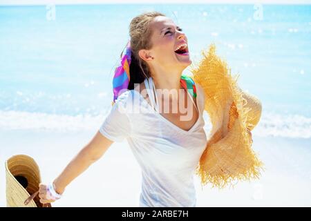 donna sorridente e moderna di mezza età in t-shirt bianca con borsa di paglia da spiaggia e grande cappello sulla riva dell'oceano. Foto Stock