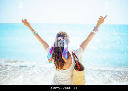 Vista da dietro alla trendy donna di 40 anni in t-shirt bianca con borsa di paglia sulla spiaggia con braccia aperte che gioisce sulla riva del mare. Foto Stock